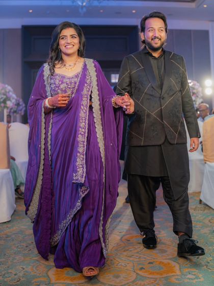 The couple walking through the aisle at their reception. The bride's flowing purple gown and the groom's modern black sherwani with geometric patterns make for a stylish and contemporary pair.