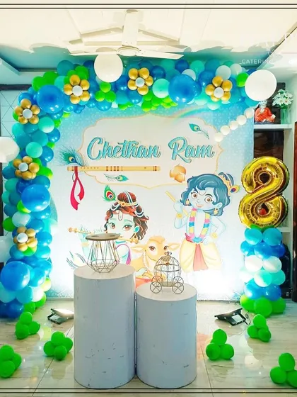 A divine Little Krishna theme for a birthday celebration. This backdrop features adorable illustrations of Krishna and Balaram, a peacock feather motif, and a blue and green balloon arch decorated with floral elements.