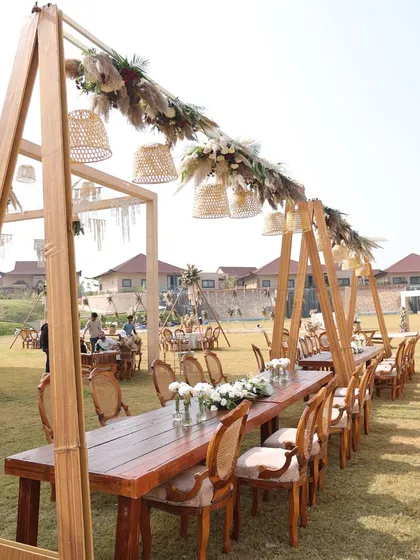 A rustic-bohemian long-table dining setup on a lawn, featuring a wooden structure overhead for hanging decor.