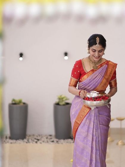 A bride carrying a silver tray during a ceremony, wearing a lavender saree with a contrasting red blouse.