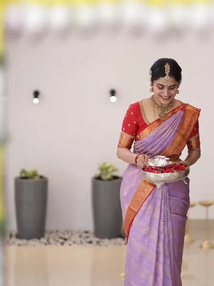 A bride carrying a silver tray during a ceremony, wearing a lavender saree with a contrasting red blouse.