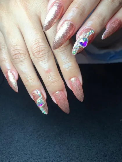 A glamorous set of long, pointed nails in a sparkling rose gold glitter finish, with large crystal accents.