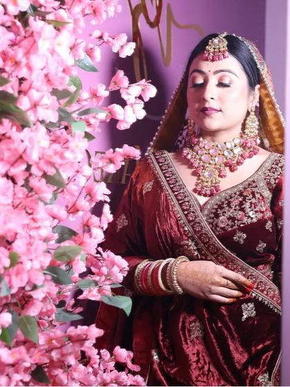 A serene bridal portrait against a backdrop of cherry blossoms. The bride wears a rich maroon velvet lehenga, complemented by a heavy Polki necklace set with pink bead drops.
