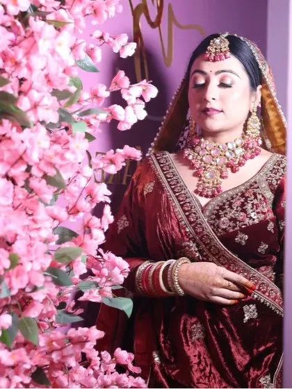 A serene bridal portrait with a backdrop of pink blossoms. The bride wears a classic maroon velvet lehenga, her look softened by the floral setting.