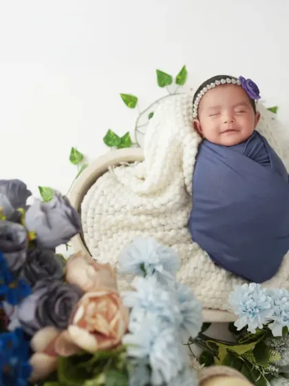This smiling newborn is swaddled in a deep blue wrap, resting in a heart-shaped basket adorned with flowers. We love capturing those fleeting, happy little moments.