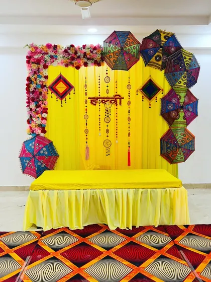 A classic Haldi backdrop with yellow drapes, the "Haldi" sign in Hindi, and colorful umbrellas. This is a popular and budget-friendly option for home ceremonies.