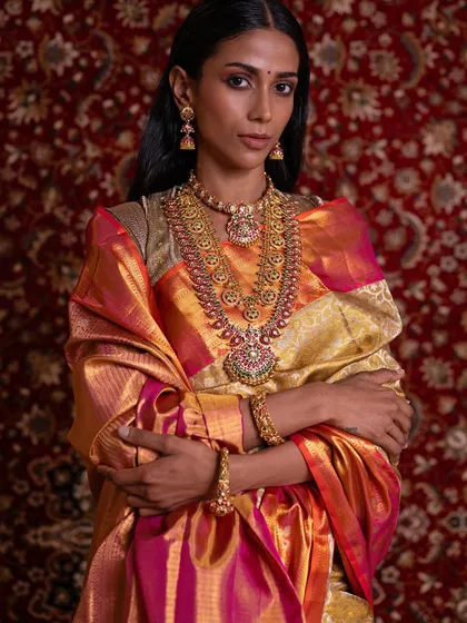 This model showcases one of our premier bridal treasures. The rich, shimmering tissue silk saree in gold and pink is paired with exquisite temple jewelry for a truly royal look.