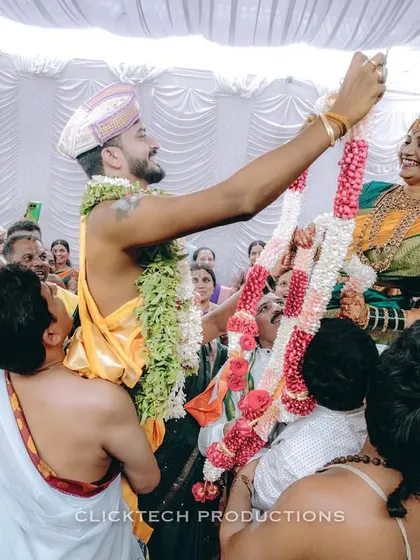 Capturing the pure joy and energy of a traditional South Indian wedding ceremony, as the groom lifts the bride during the garland exchange ritual, surrounded by smiling family.