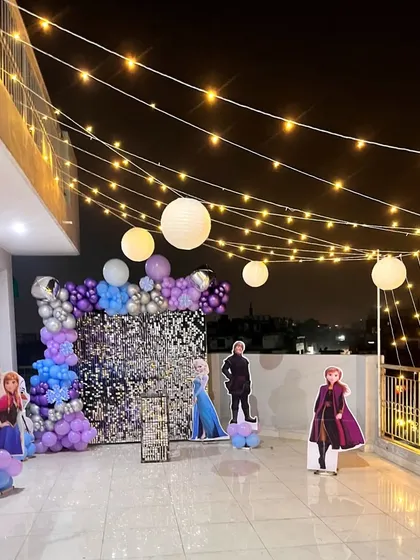 A 'Frozen' themed birthday party on a rooftop terrace at night. The string lights, paper lanterns, and illuminated backdrop create a magical winter wonderland.