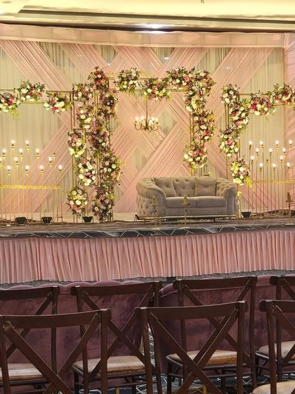 This wide shot shows a complete reception stage design before the event. The structure includes a metal framework adorned with flowers, a soft grey couple's sofa, and candle-like lighting against a draped backdrop.
