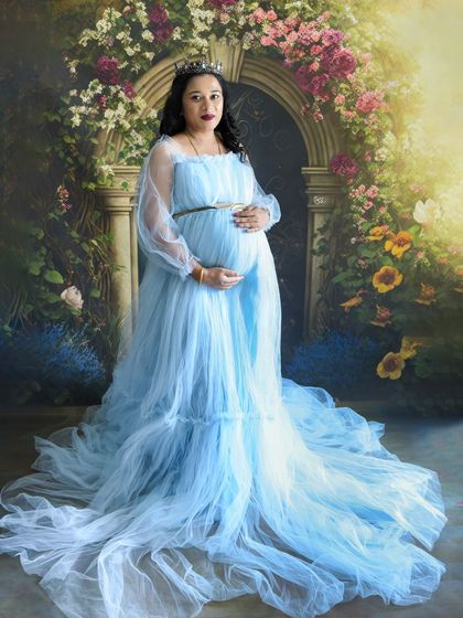 A full-length portrait of a mom-to-be in a flowing light blue gown and tiara, looking like a queen in our studio.