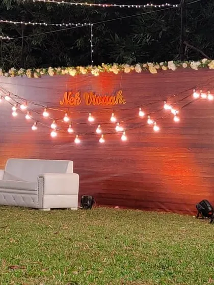 For a warm and intimate feel, we designed this rustic engagement backdrop. It features a rich wooden panel, a personalized neon sign with the couple's name, and strings of cozy Edison bulbs, perfect for an outdoor lawn party.