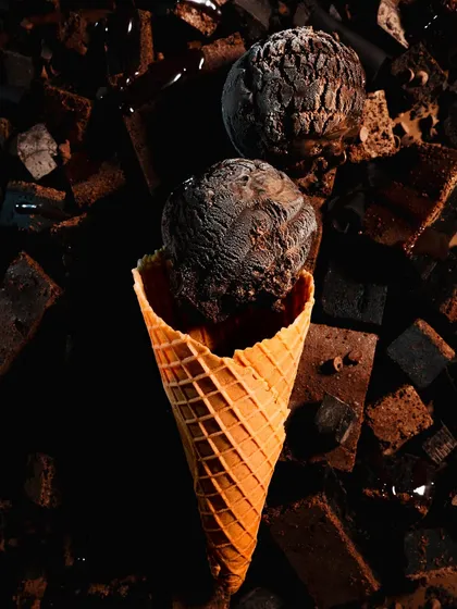 A dark and moody shot of chocolate ice cream on a cone, surrounded by brownie pieces. The dramatic lighting highlights the rich texture of the ice cream.