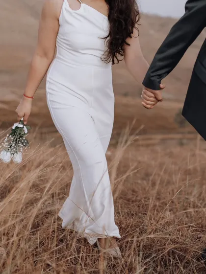 A close-up detail shot of the couple holding hands as they walk, with the bride holding a simple bouquet of white roses. A symbol of their shared path.