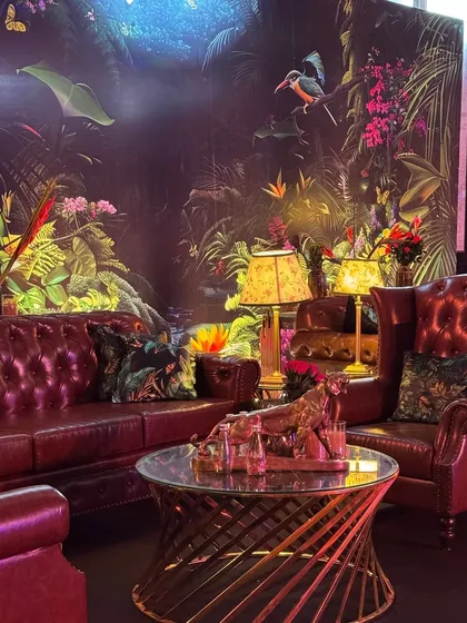 A luxurious lounge corner from the "Jungle" themed party. A classic leather chesterfield sofa is set against a vibrant, tropical-print wallpaper, creating a moody and sophisticated vibe.