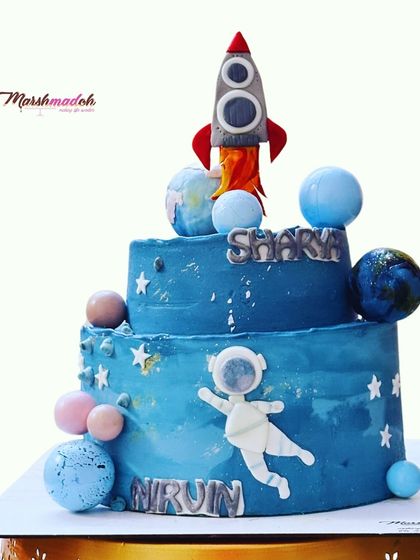 A two-tiered space and astronaut theme cake perfect for a young explorer's birthday. The cake is decorated with planets, stars, a rocket, and an astronaut, all floating in a galaxy of blue buttercream.