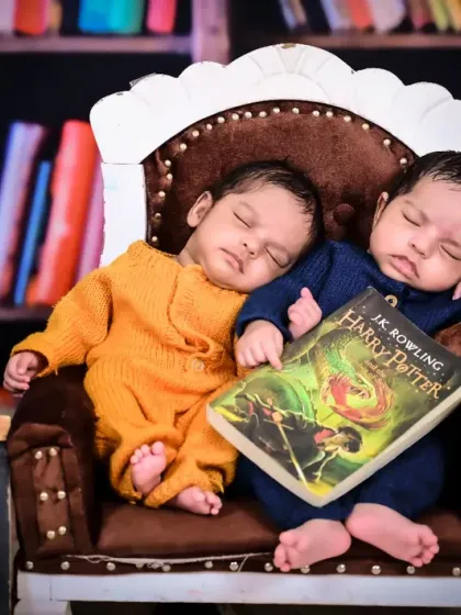 Twin newborns sleeping in a chair in a library setup, one holding a Harry Potter book.