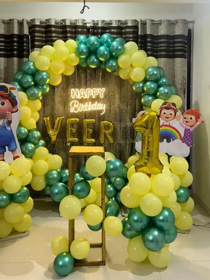 A Cocomelon first birthday decoration for a home setting. This setup includes a circular balloon arch in green and yellow, a "Happy Birthday" neon sign, and character cutouts.