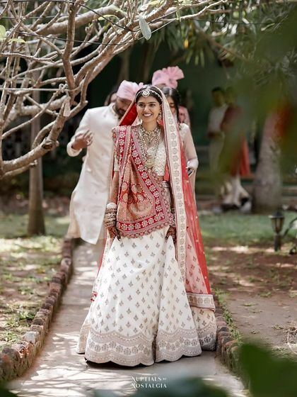A bride makes her entrance, walking down a garden path, her white and red lehenga radiant in the sunlight.