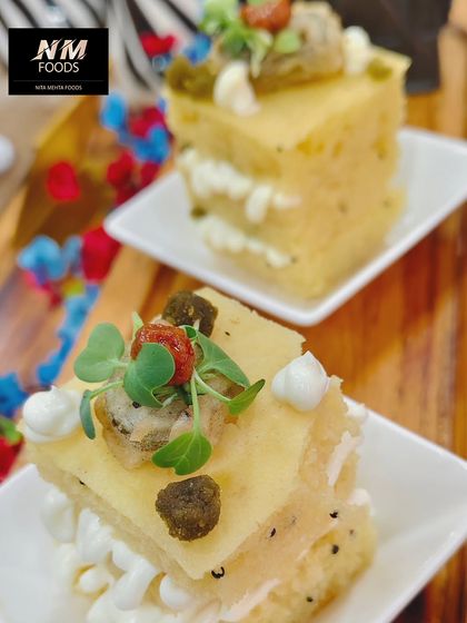 A savory cake-like appetizer, featuring layers of fluffy dhokla with a creamy filling and garnished with microgreens and sun-dried tomatoes. A unique and elegant bite.