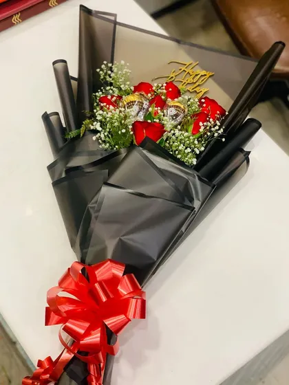 A birthday-themed bouquet featuring red roses, baby's breath, and assorted chocolates. The arrangement is topped with a golden "Happy Birthday" sign and wrapped in sleek black paper.