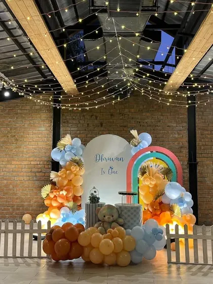 A cute teddy bear theme decoration inside a venue with a rustic brick wall. The setup includes a small white picket fence, a rainbow arch, and a color palette of orange and light blue balloons.