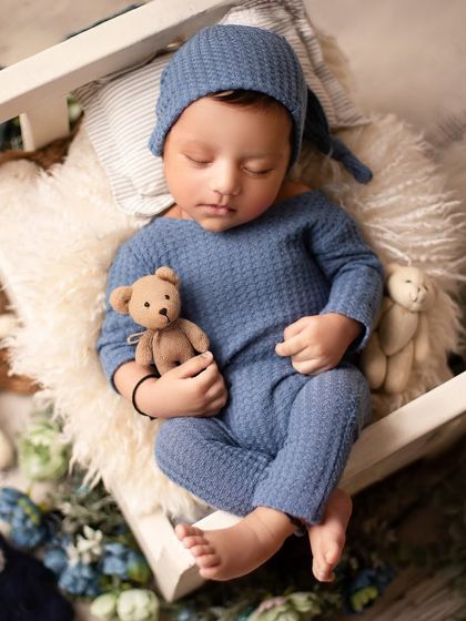 A sweet baby boy in a blue knit outfit, cuddling his teddy bear on a miniature bed. This setup creates a classic, storybook feel.