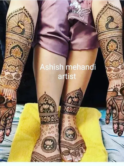 A classic bridal pose showing full coverage mehandi on both hands and feet, featuring lotus and peacock motifs.