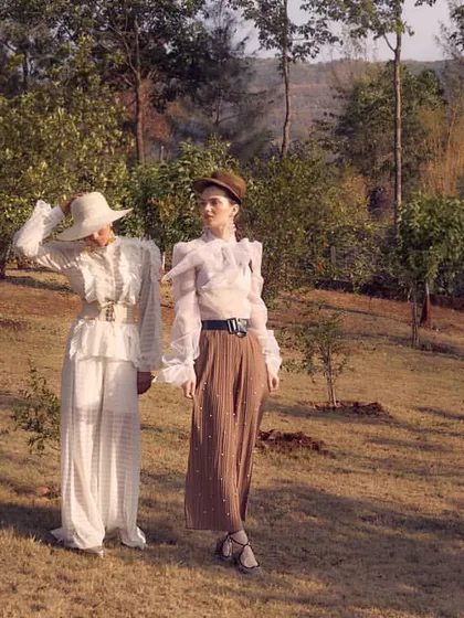 A two-model shot for Starch Label's "White Rose" collection, set in a sunlit field. The styling combines different textures and silhouettes, like a sheer blouse with trousers and a full white dress, to create a cohesive, vintage-inspired story.