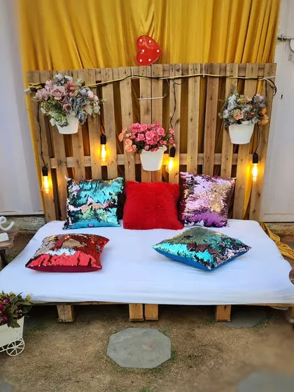 A simple and cozy seating area created with a wooden pallet base. It's decorated with fun, reversible sequin pillows and hanging flower pots against a rustic pallet wall, perfect for a casual photo booth or lounge spot.