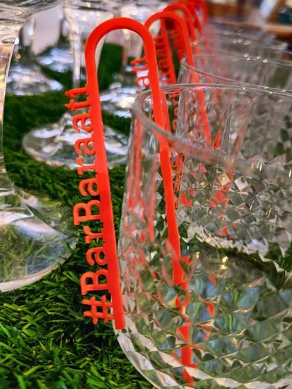 A close-up of a custom-made, bright red drink stirrer with the '#BarBaraati' hashtag, designed to sit perfectly on the rim of a crystal glass. It's a small detail that adds a big pop of color and branding.