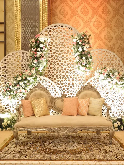 An elegant stage setup with a vintage-style sofa, set against a white jaali-work backdrop adorned with delicate pink and white floral arrangements.