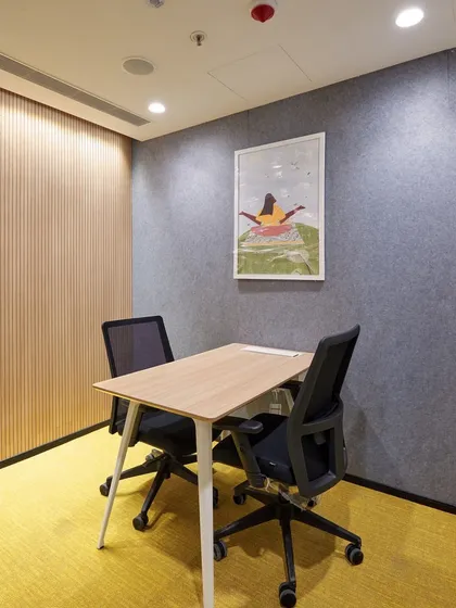 A small, two-person meeting room, perfect for one-on-one conversations or focused work. The yellow carpet and simple artwork add a touch of warmth.