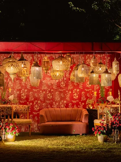 An intimate seating area for a traditional ceremony. The backdrop features a rich red floral-print fabric and a collection of antique-style hanging lanterns, creating a warm and soulful corner.