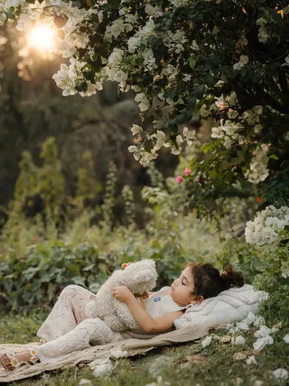 A young girl sleeps peacefully on a blanket under a flowering tree, cuddling a teddy bear. This serene portrait is like a scene from a fairytale.