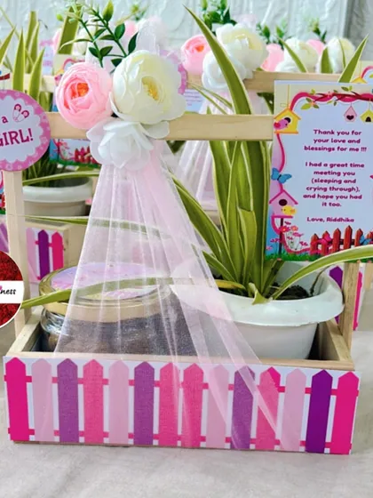A charming "It's a Girl!" announcement favor, featuring a pink picket fence design, a live plant, and a jar of cookies, complete with a sweet thank you note.