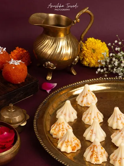 A tray of white modaks and orange ladoos, styled with a brass pitcher and flowers. The composition feels like a snapshot from a real festive prayer ceremony.