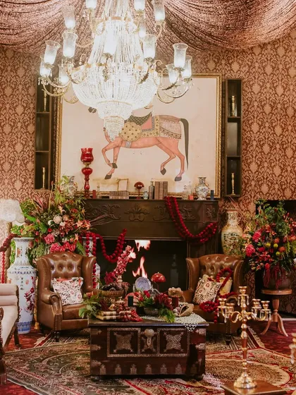 A cozy corner arranged around a fireplace, featuring classic leather armchairs, a large traditional Indian painting, and rich red floral arrangements. This vignette was designed to feel like a room in a grand, old-world mansion.