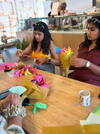 Two participants work together at a table filled with colorful paper and finished origami flowers. Our workshops foster a collaborative spirit, where people help each other and share in the creative process.