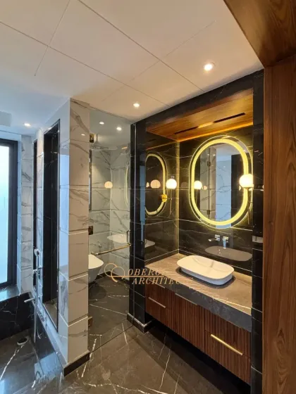 A dramatic and sophisticated bathroom design featuring black marble walls and floors, contrasted with warm wood accents. The backlit oval mirror adds a modern, glamorous touch.