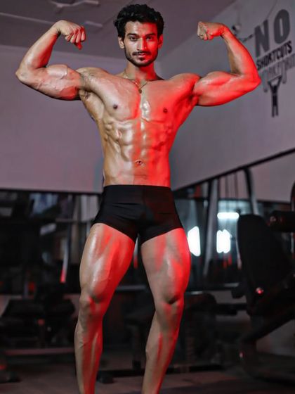 A front double biceps pose under red lighting. This pose is a classic display of arm size, symmetry, and a tapered waist.