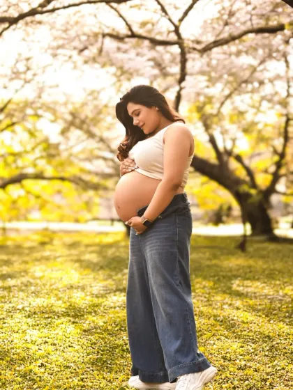 A casual and beautiful outdoor maternity portrait. The expecting mother cradles her bare bump against a backdrop of lush, sunlit trees.