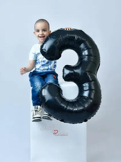 A happy three year old boy celebrating his birthday in the studio. He is sitting on a white block next to a large black balloon in the shape of the number three.