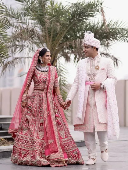 A candid shot of a newly married couple walking hand-in-hand, showcasing the bride's vibrant pink lehenga and the groom's elegant sherwani in a natural, outdoor setting.
