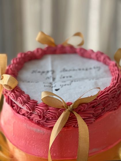 A romantic burn away cake in pink with gold ribbons, perfect for an anniversary. The top layer features a handwritten message, adding to the personal feel.