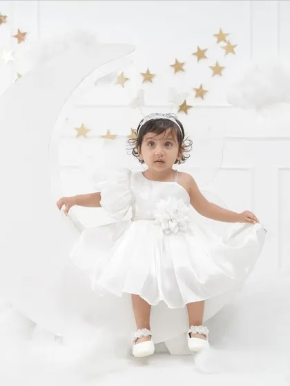 A whimsical 'Moon & Stars' themed portrait of a little girl in a beautiful white dress, sitting on a large crescent moon prop surrounded by clouds and stars.