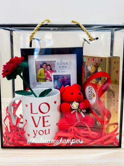 This Valentine's hamper comes in a modern acrylic tote bag. It's filled with a "Love You" mug, a personalized photo frame, a handmade crochet rose, a teddy, and Ferrero Rochers.