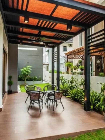 This covered patio design features a dark metal frame pergola with a contrasting orange fabric roof, providing shade and a pop of color. It's an ideal setup for an outdoor dining area, complete with comfortable seating and surrounding greenery.