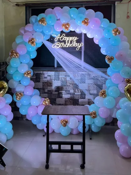 A simple yet beautiful balloon arch decoration perfect for a small home celebration. The pastel blue, pink, and gold balloons create a soft and dreamy look, centered around a classic 'Happy Birthday' neon sign.