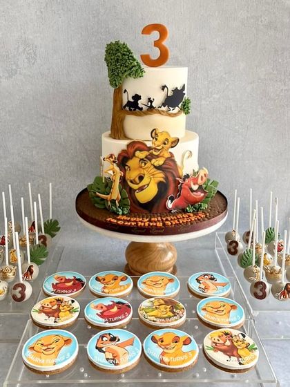 A full dessert table for a Lion King fan! The two-tiered cake features hand-painted scenes from the movie, and it's surrounded by themed cookies and cake pops with all the beloved characters.